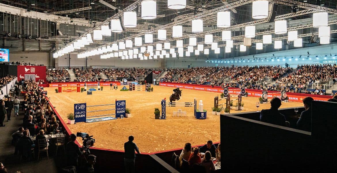 Espectáculo de salto ecuestre en pabellón cubierto con espectadores disfrutando del evento en un entorno moderno y bien iluminado.