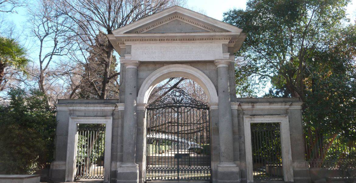 Fachada del Parque del Retiro en Madrid, entrada neoclásica con puertas de hierro y columnas, rodeada de vegetación, buen estado, lugar histórico y cultural.