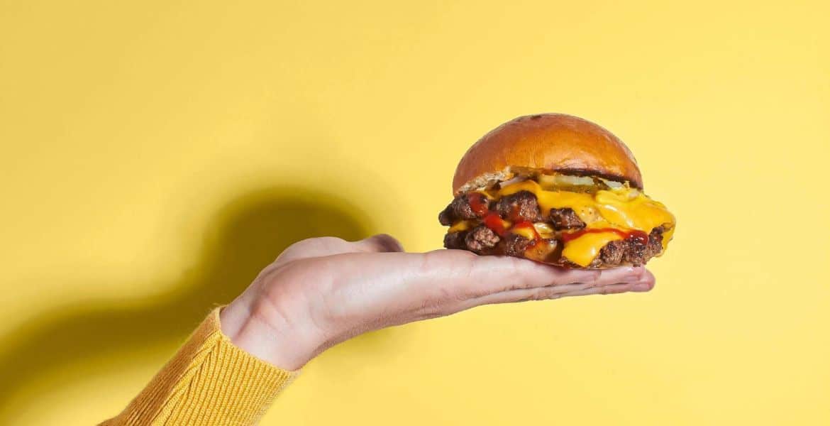 Hamburguesa doble con queso, carne jugosa, cebolla y salsas en un pan dorado, en un fondo amarillo brillante, ilustrando comida rápida y personalizada en Hotel Madrid Río.