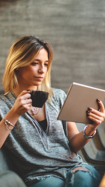 Una mujer leyendo en un café, disfrutando de su tiempo libre, con taza en mano y usando una tableta. Ambiente moderno y relajante.