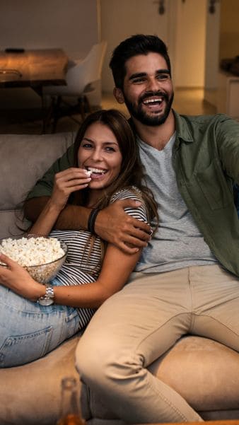 Pareja feliz disfrutando en casa, viendo televisión y comiendo palomitas, concepto de ocio y relajación en un entorno doméstico cálido y acogedor.