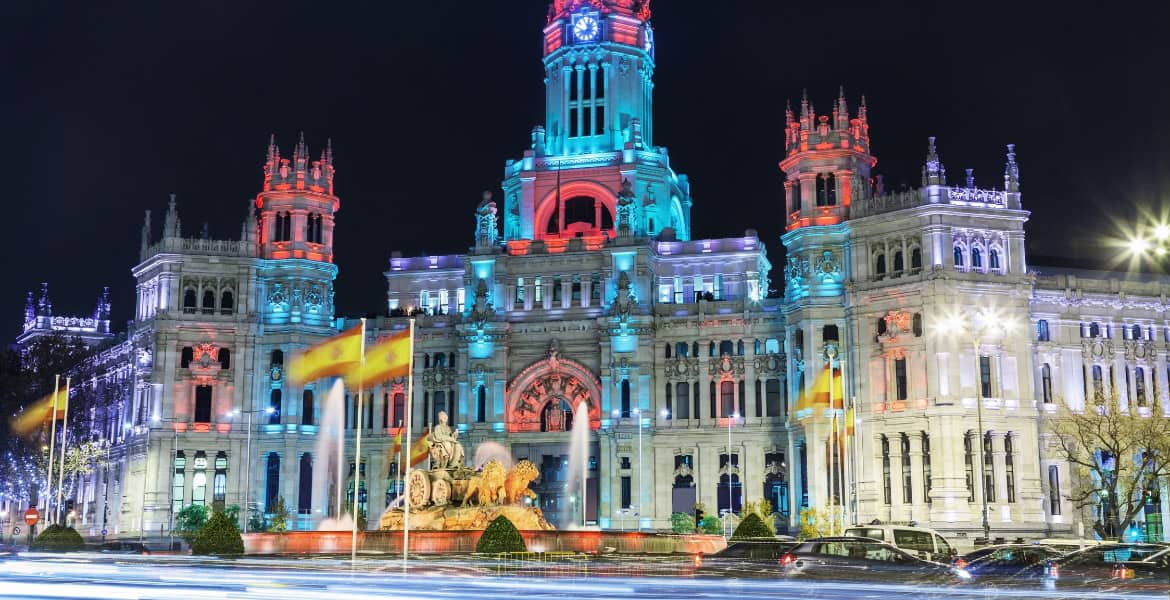 Imponente Ayuntamiento de Madrid iluminado con luces de colores en la noche, cerca del río Manzanares, en un entorno urbano emblemático y turístico.