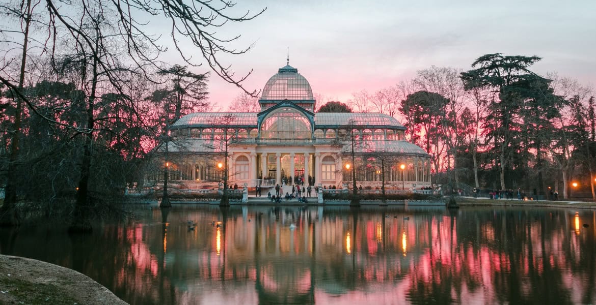 Cúpula de cristal en el Palacio de Cristal del Parque del Retiro, Madrid, al atardecer con reflejo en el estanque cercano.