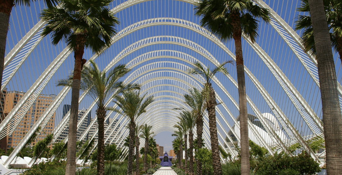 Estructura moderna de vidrio y acero en el Parque Madrid Río, con palmeras y paisaje urbano de Madrid.