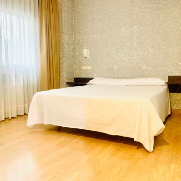 Cama doble en habitación de hotel con decoración moderna y ventanas grandes con cortinas, ideal para estancias en Madrid Río.
